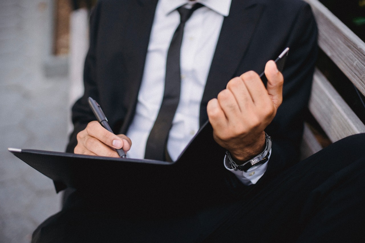 A main in a black suit and white shirt writes with pen in the black notebook, showcasing black wristwatch