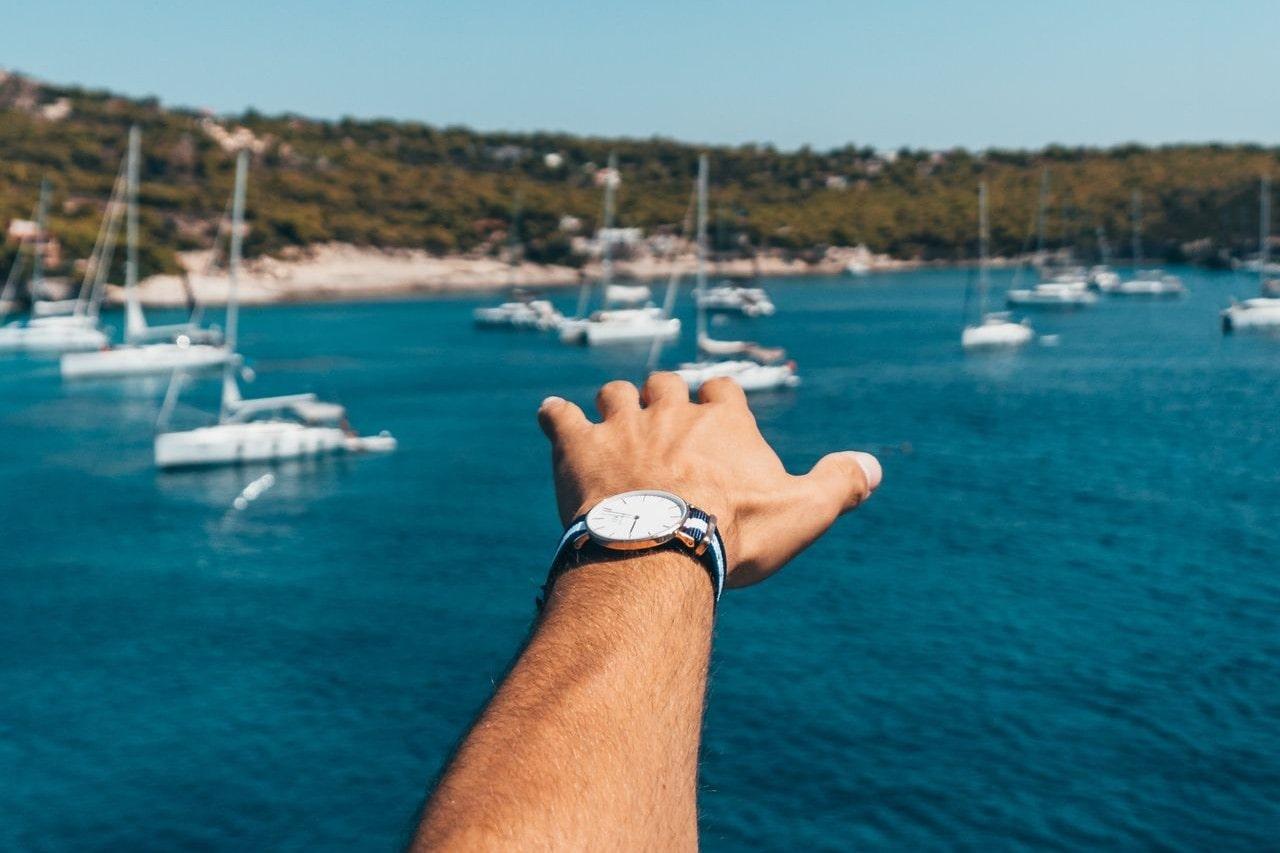A close-up of a man’s hand wearing a luxury watch, reaching out toward the water filled with sailboats.