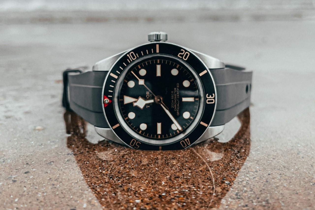 A close-up of a black, gold, white, and silver TUDOR watch sitting in shallow water on a granite surface.