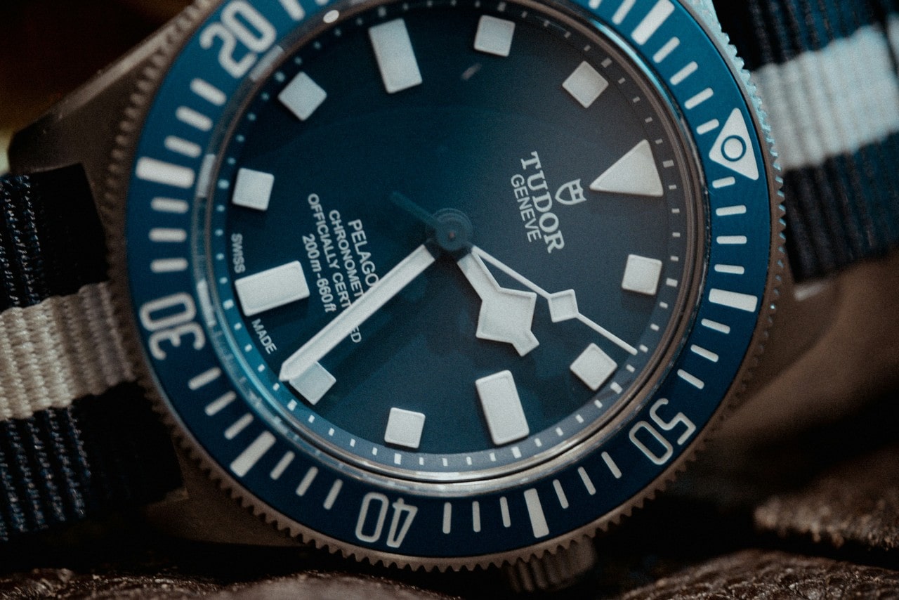 A close-up image of a silver and blue TUDOR watch with a blue and white striped strap.