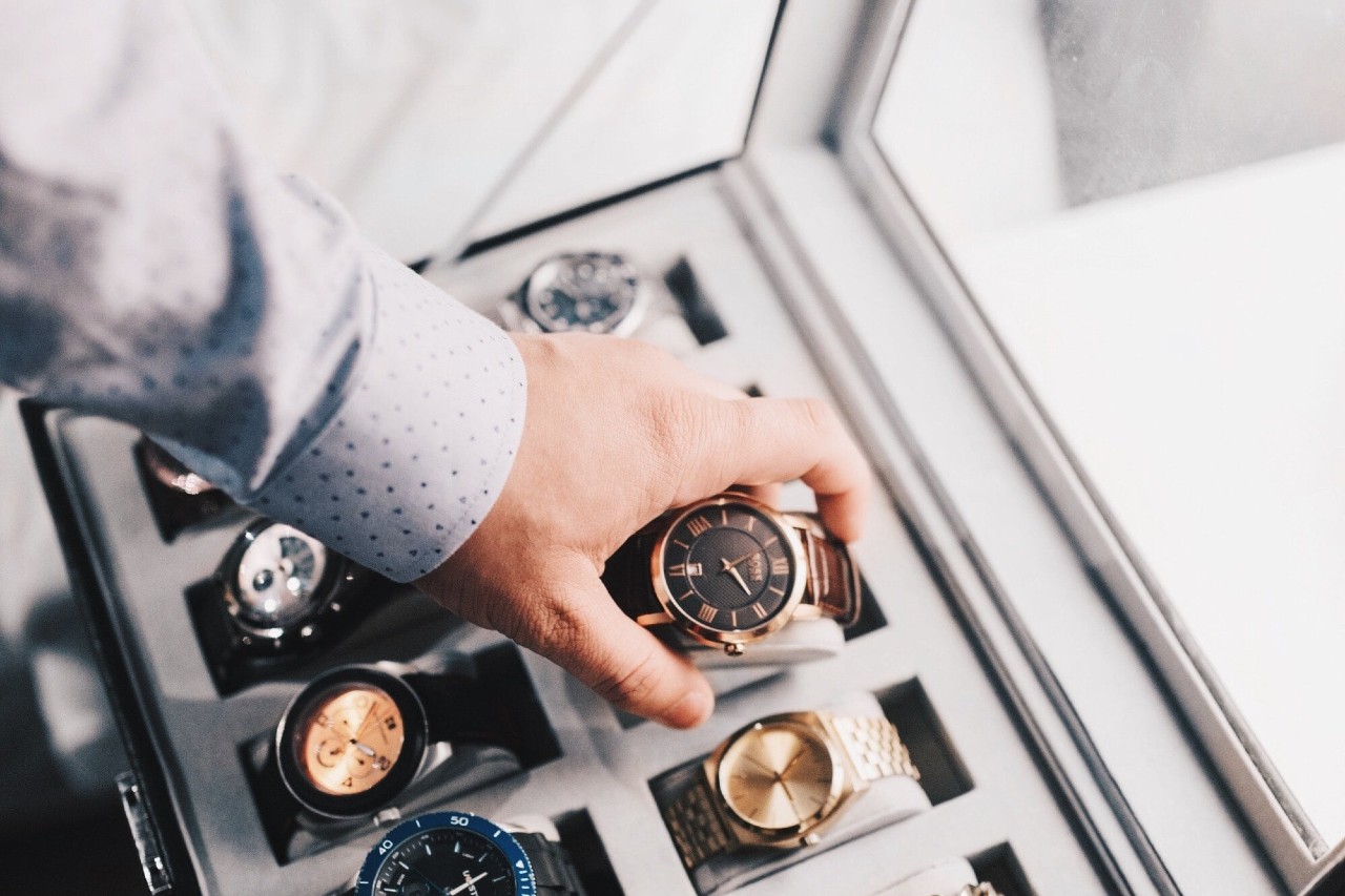 A man’s hand selecting a brown leather wristwatch from a display case filled with multiple luxury timepieces. A man’s hand selecting a brown leather wristwatch from a display case filled with multiple luxury timepieces.